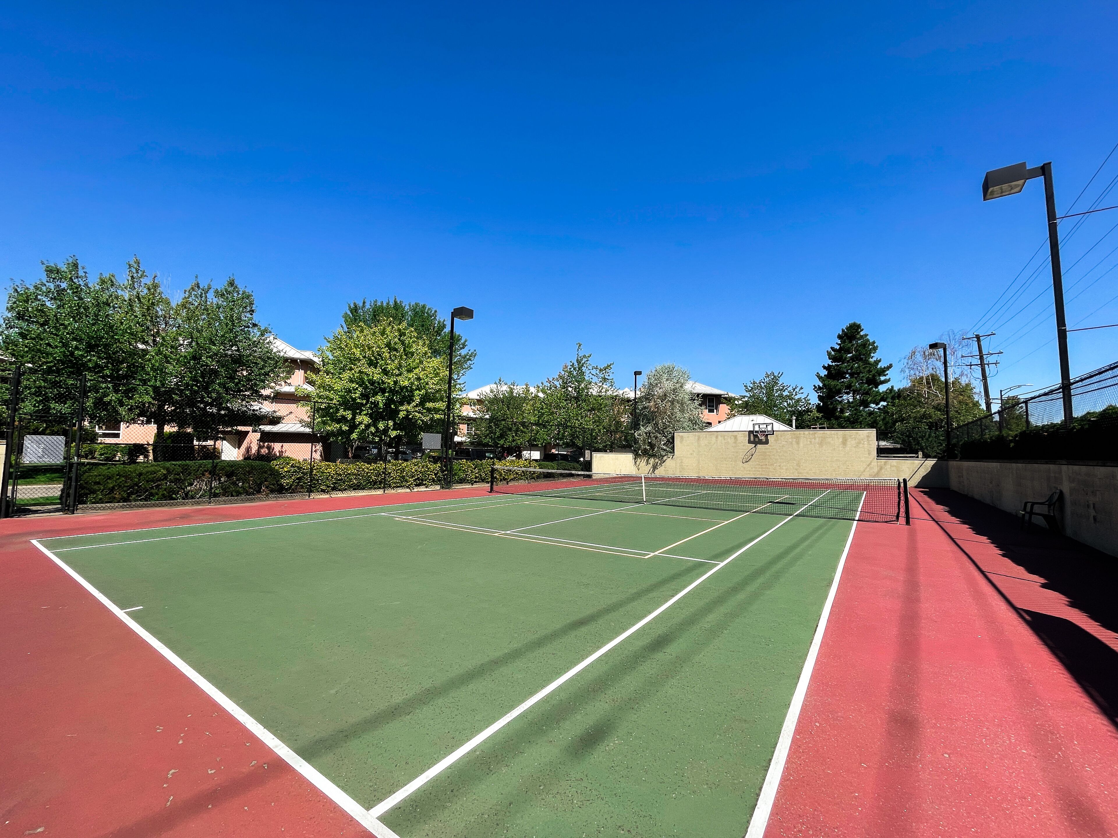 A tennis court with a red and green surface, surrounded by trees and buildings.