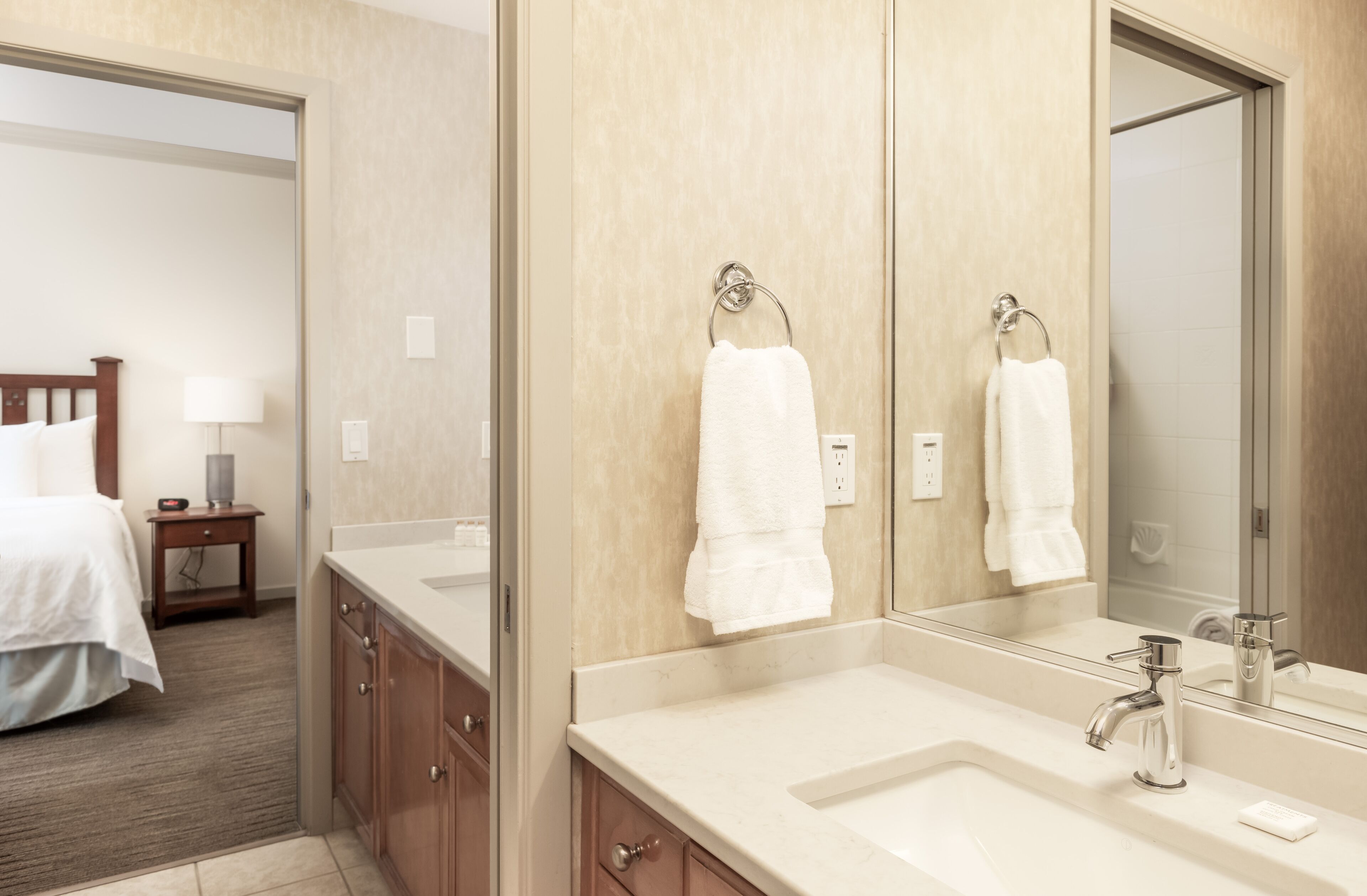 A hotel bathroom with a large mirror, two sinks, a wooden cabinet, and a bed in the adjacent room.