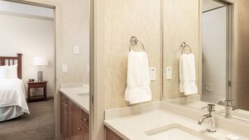 A hotel bathroom with a large mirror, two sinks, a wooden cabinet, and a bed in the adjacent room.