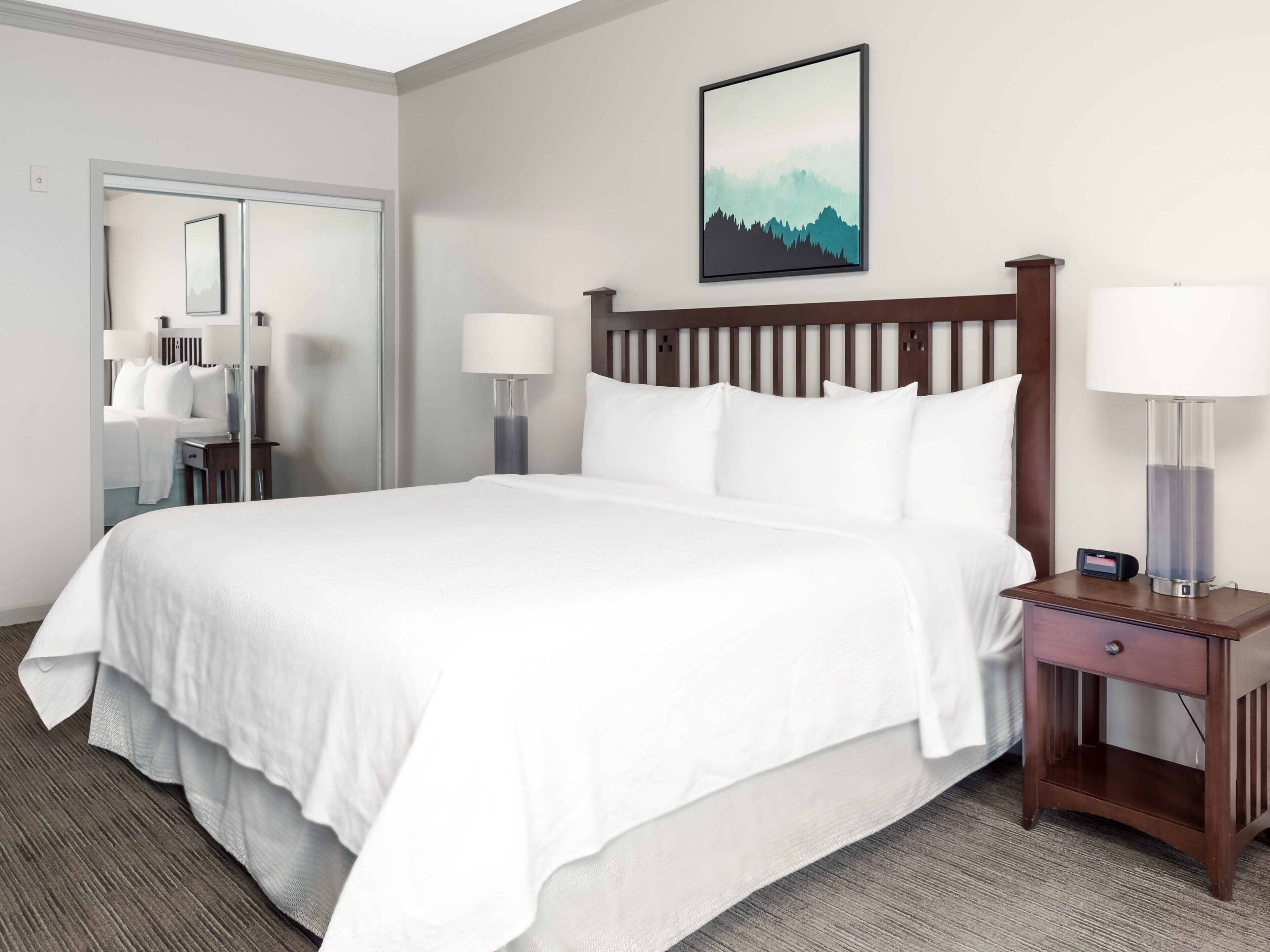 A neatly made bed with white linens, a wooden headboard, two bedside lamps, and a nightstand with a lamp and a small electronic device.