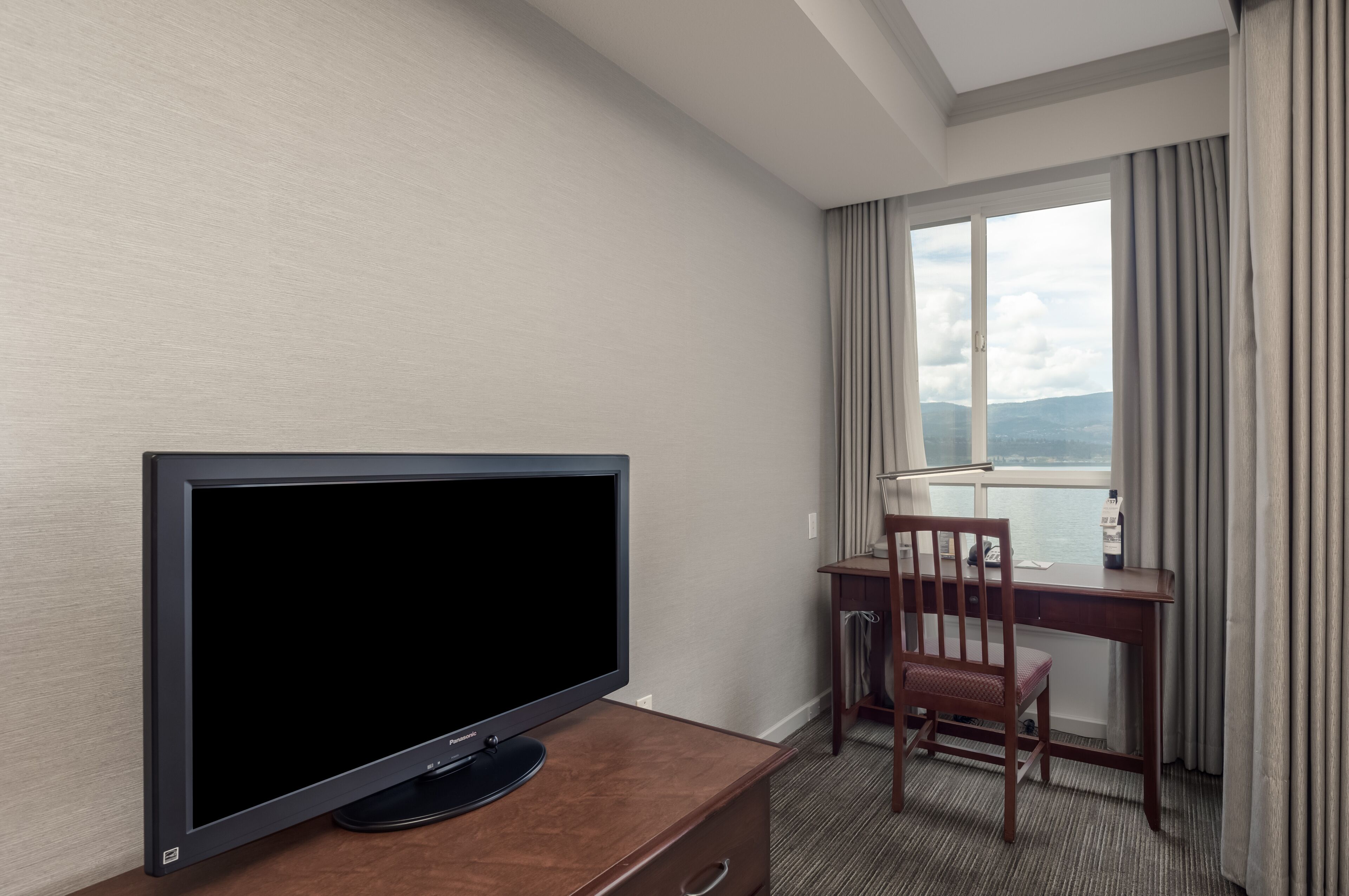 Ett hotellrum med en platt-TV placerad på en byrå i trä, ett skrivbord med en stol och ett stort fönster med gardiner.