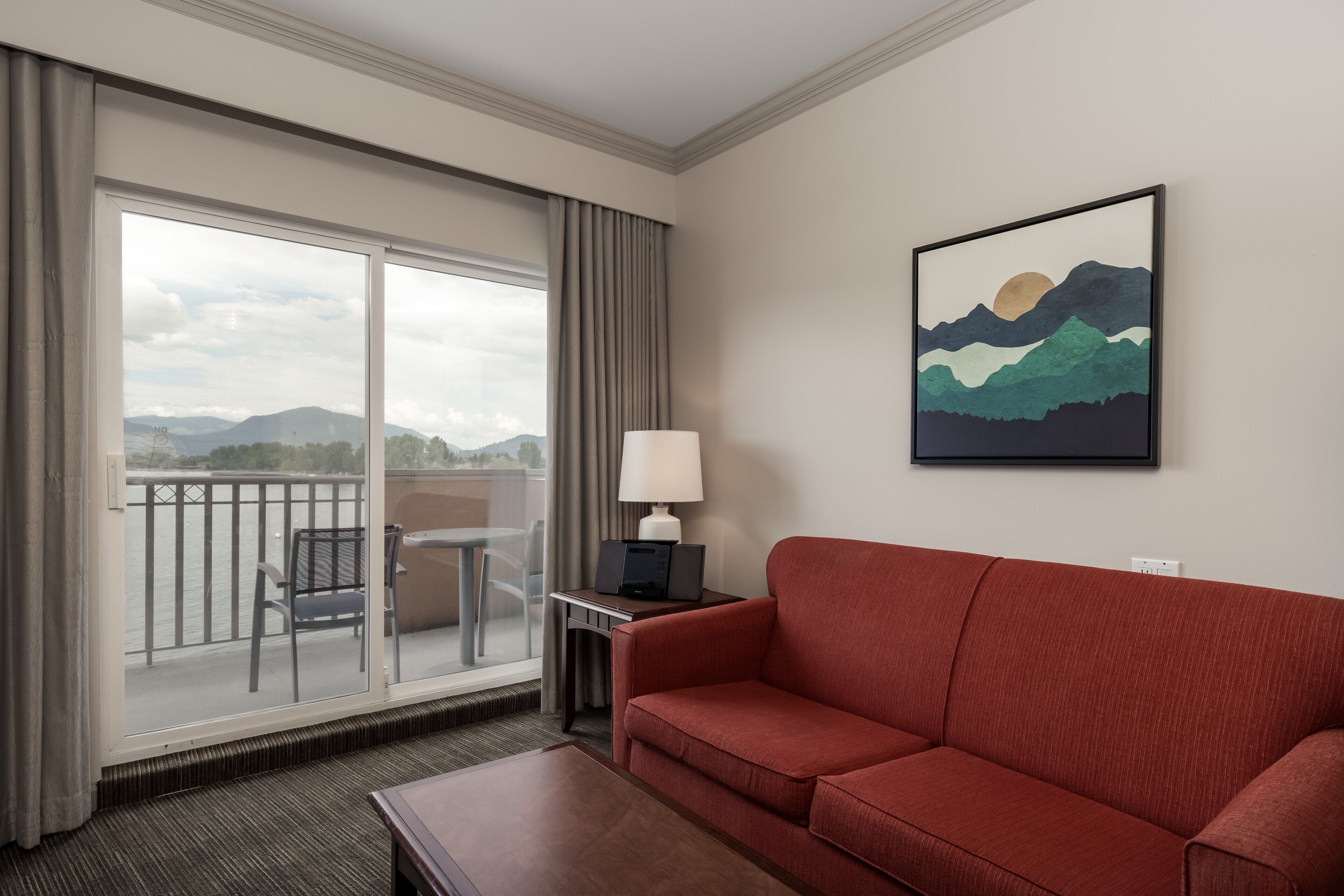 A hotel room with a red sofa, a desk with a lamp, and a large window offering a view of mountains and a body of water.