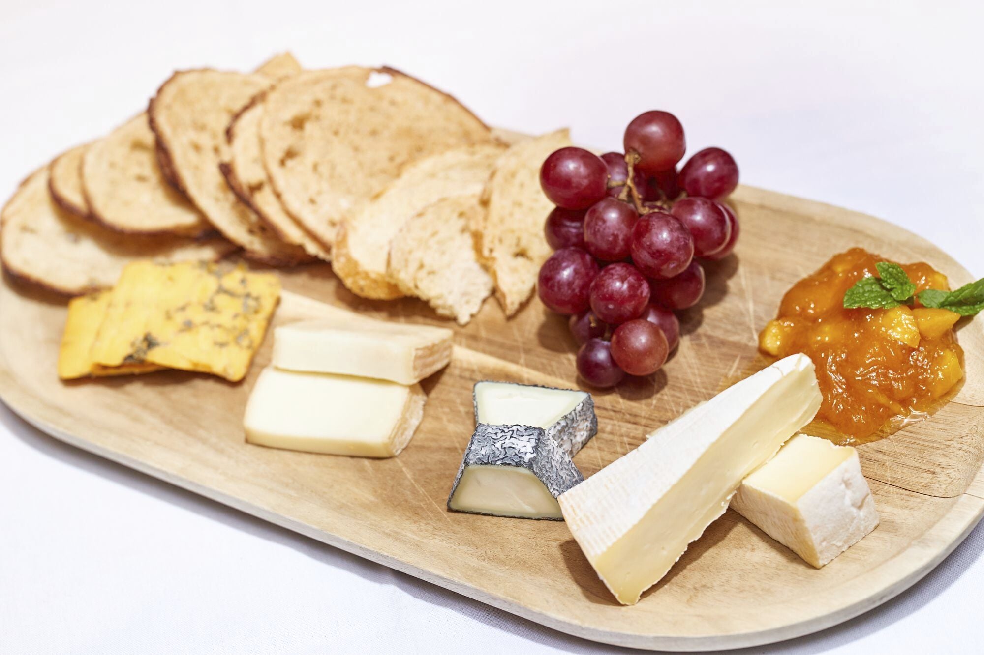 A wooden board with various cheeses, slices of bread, grapes, and a fruit preserve.