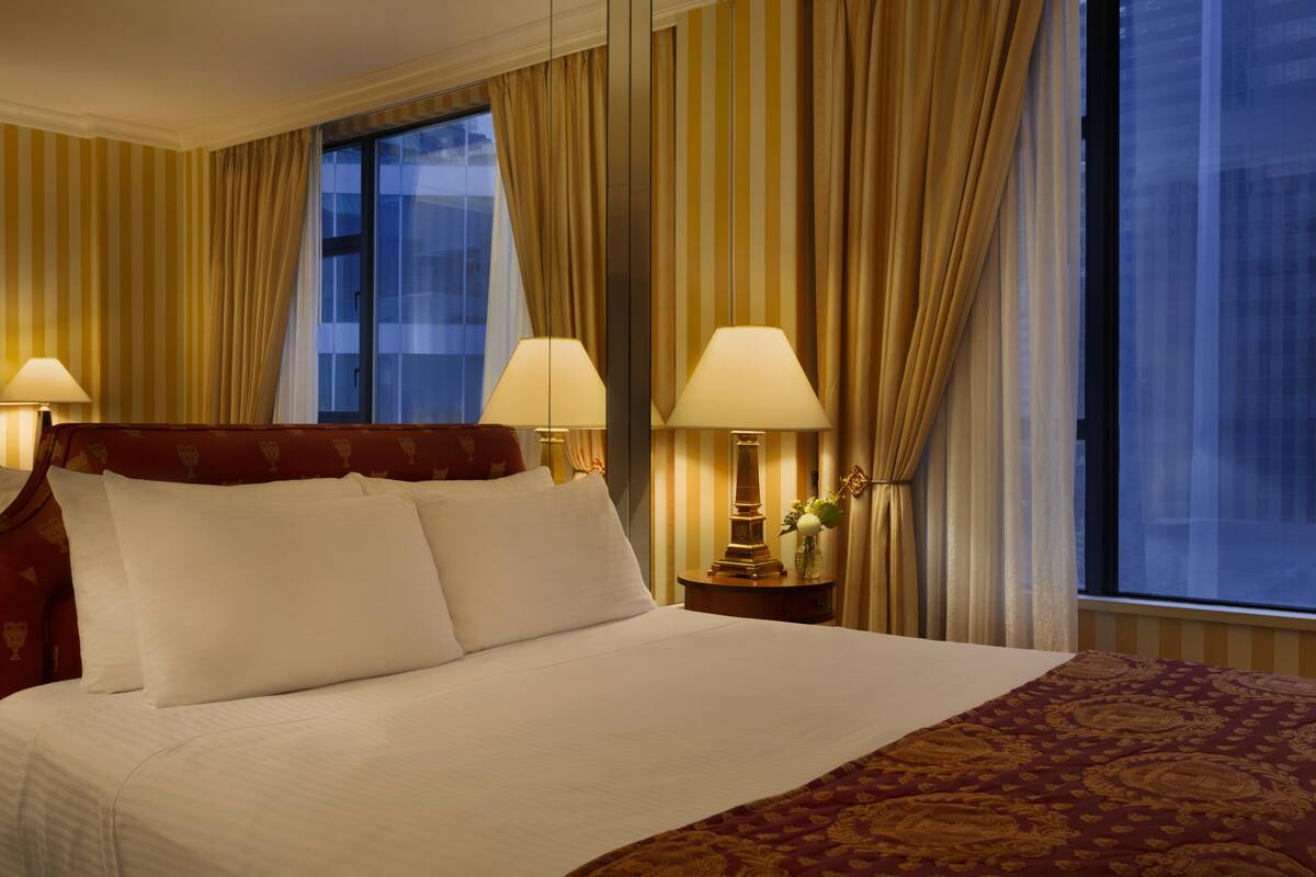 A hotel room with a large bed, two bedside lamps, a nightstand with a vase of flowers, and a window with curtains.