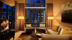 A modern hotel room with a large window, two armchairs, a sofa, a coffee table, and a flat-screen TV.