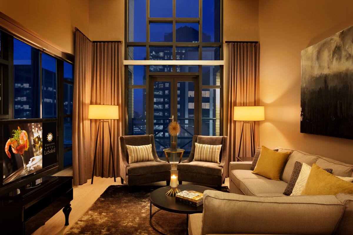 A modern hotel room with a large window, two armchairs, a sofa, a coffee table, and a flat-screen TV.