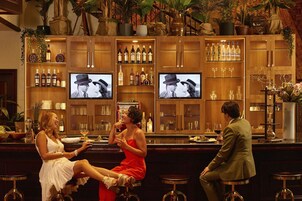 Zona de bar con múltiples estantes llenos de botellas, dos televisores mostrando imágenes y diversos taburetes.