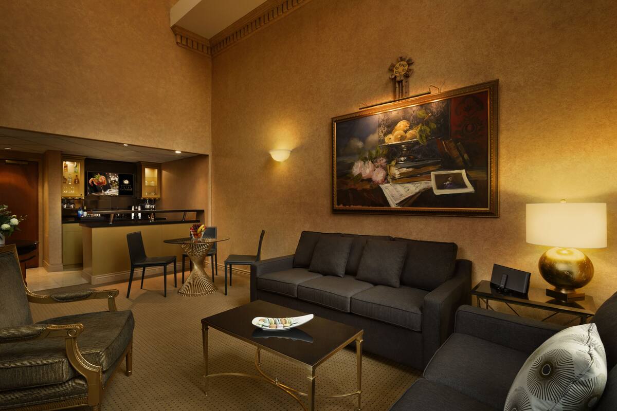 A hotel lobby with a sofa, armchairs, a coffee table, a dining area with chairs, a bar, and a painting on the wall.