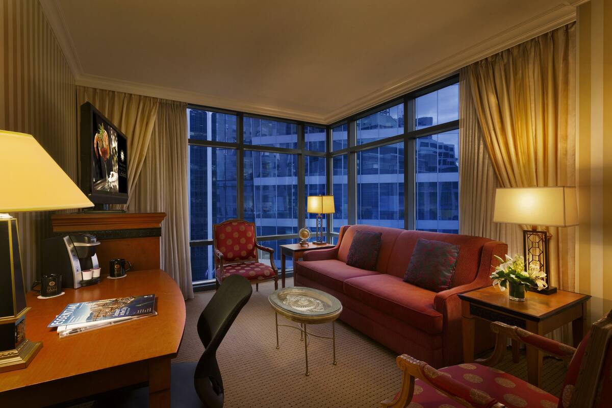 A hotel room with a large window, a red sofa, a small table, a desk with a lamp, and a chair.