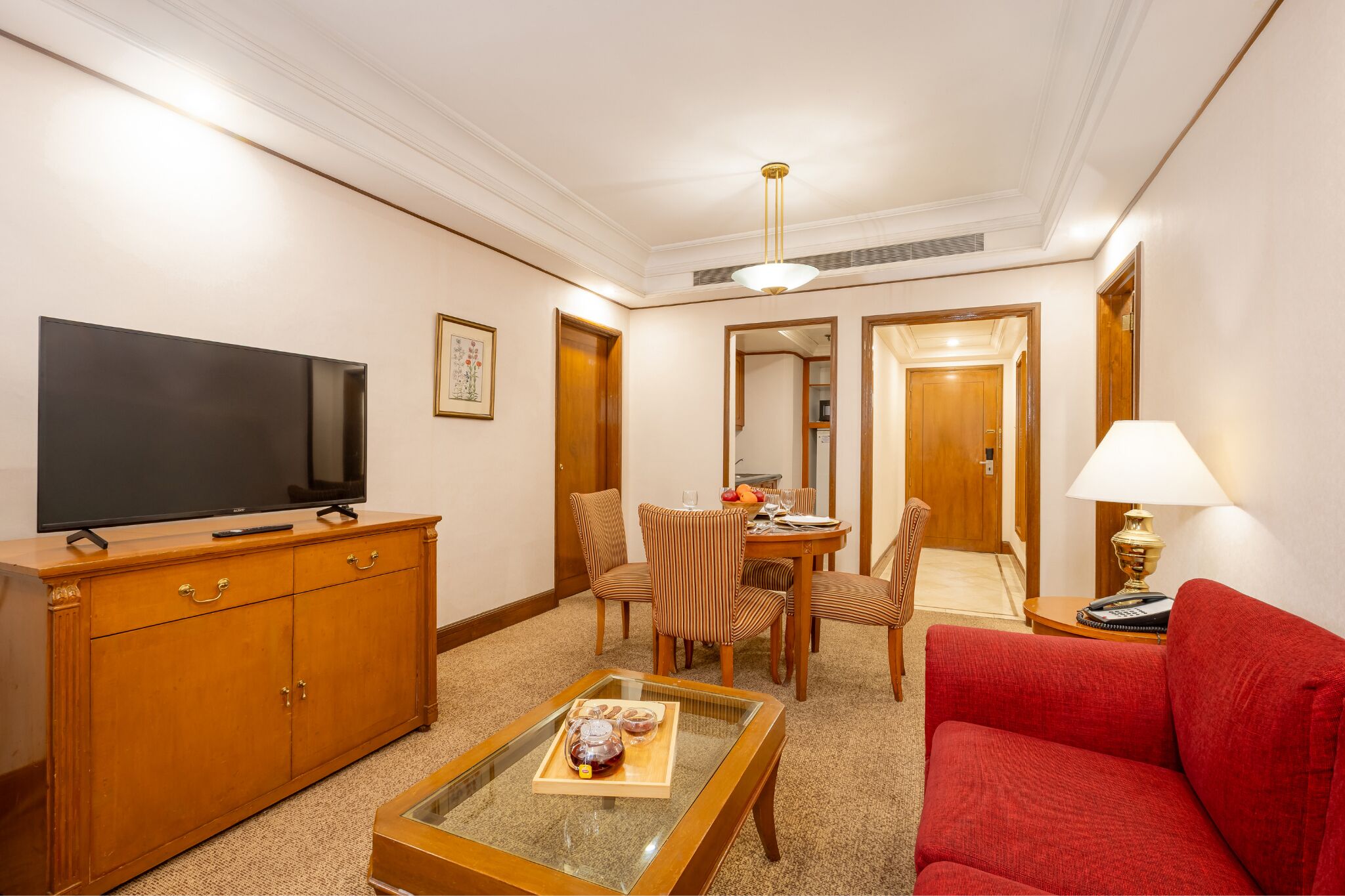Quarto de hotel com televisão de tela plana, móveis de madeira, mesa de jantar com cadeiras, sofá vermelho e luminária.