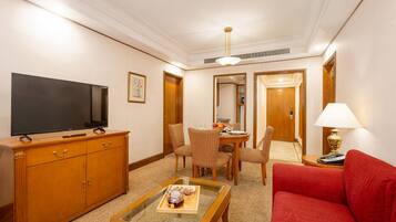 Quarto de hotel com televisão de tela plana, móveis de madeira, mesa de jantar com cadeiras, sofá vermelho e luminária.