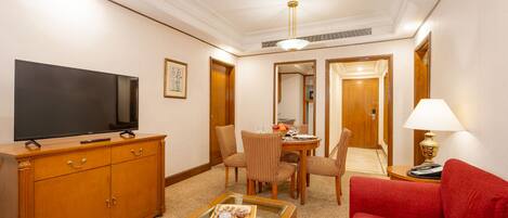 A hotel room with a flat-screen TV, wooden furniture, a dining table with chairs, a red sofa, and a lamp.