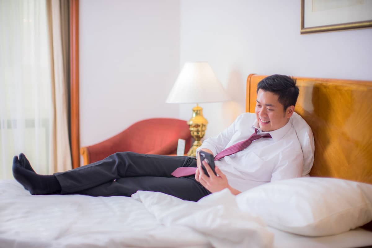 A person lying on a bed in a hotel room with a lamp and a framed picture on the wall.