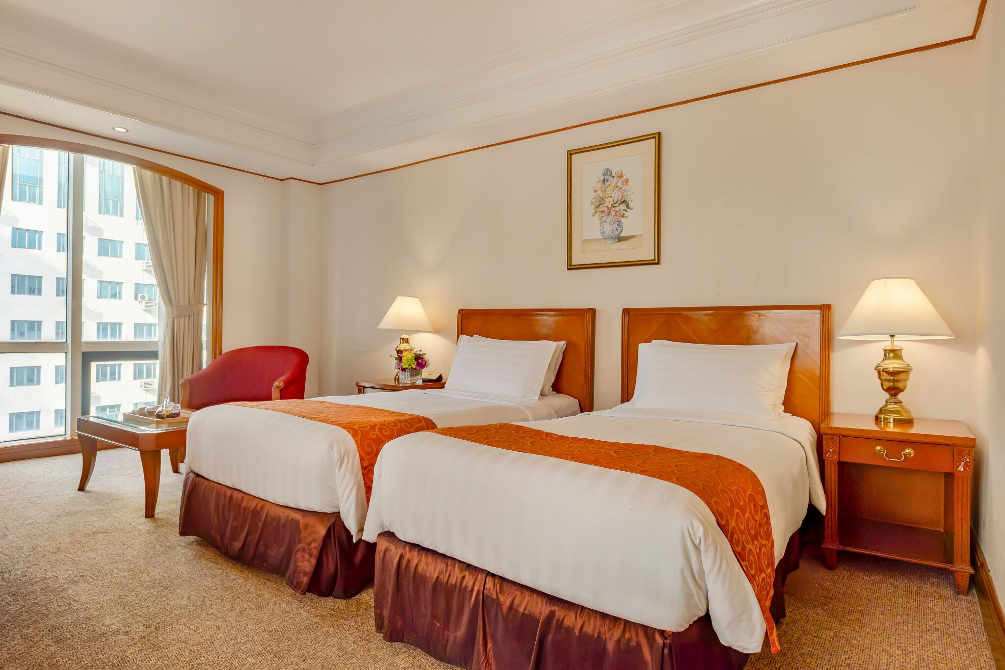 Une chambre d’hôtel avec deux lits, une chaise rouge, une petite table avec une lampe et une fenêtre avec des rideaux.