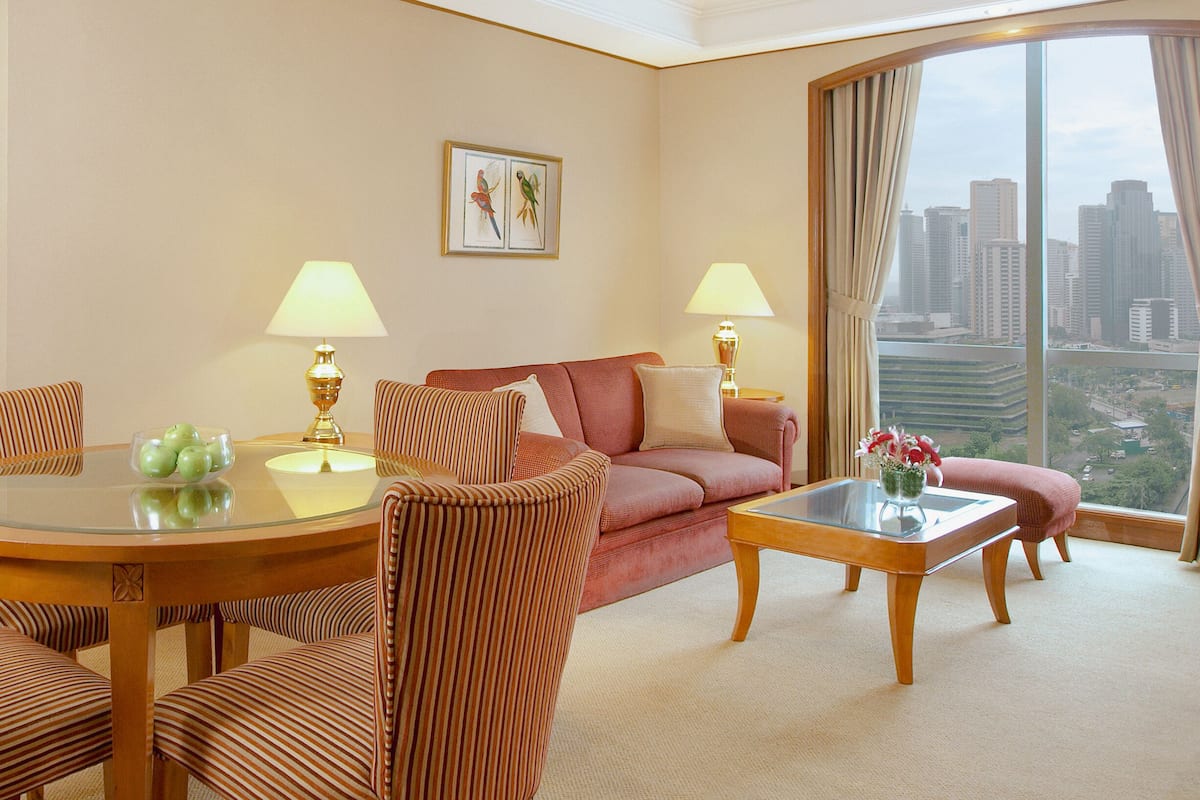A hotel room with a sofa, armchairs, a dining table, and a large window with a city view.