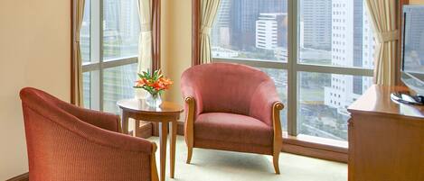 A hotel room with two armchairs, a small table with a vase of flowers, and a desk with a television. Large windows offer a city view.