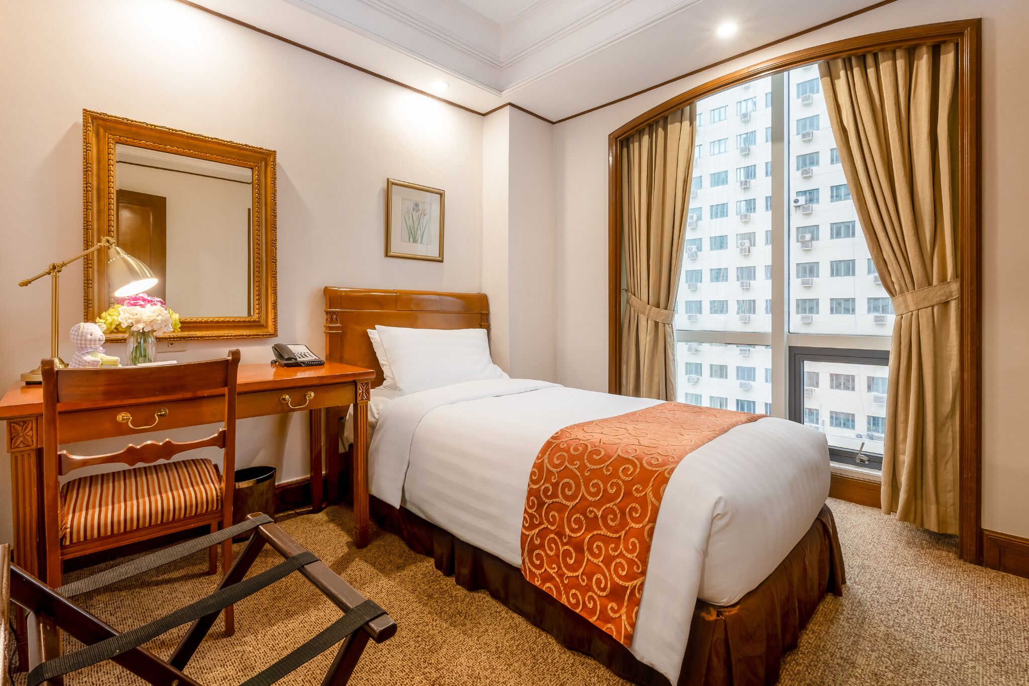 Une chambre d’hôtel avec un lit, un bureau doté d’une lampe, une chaise, une fenêtre avec des rideaux et une vue sur les bâtiments environnants.