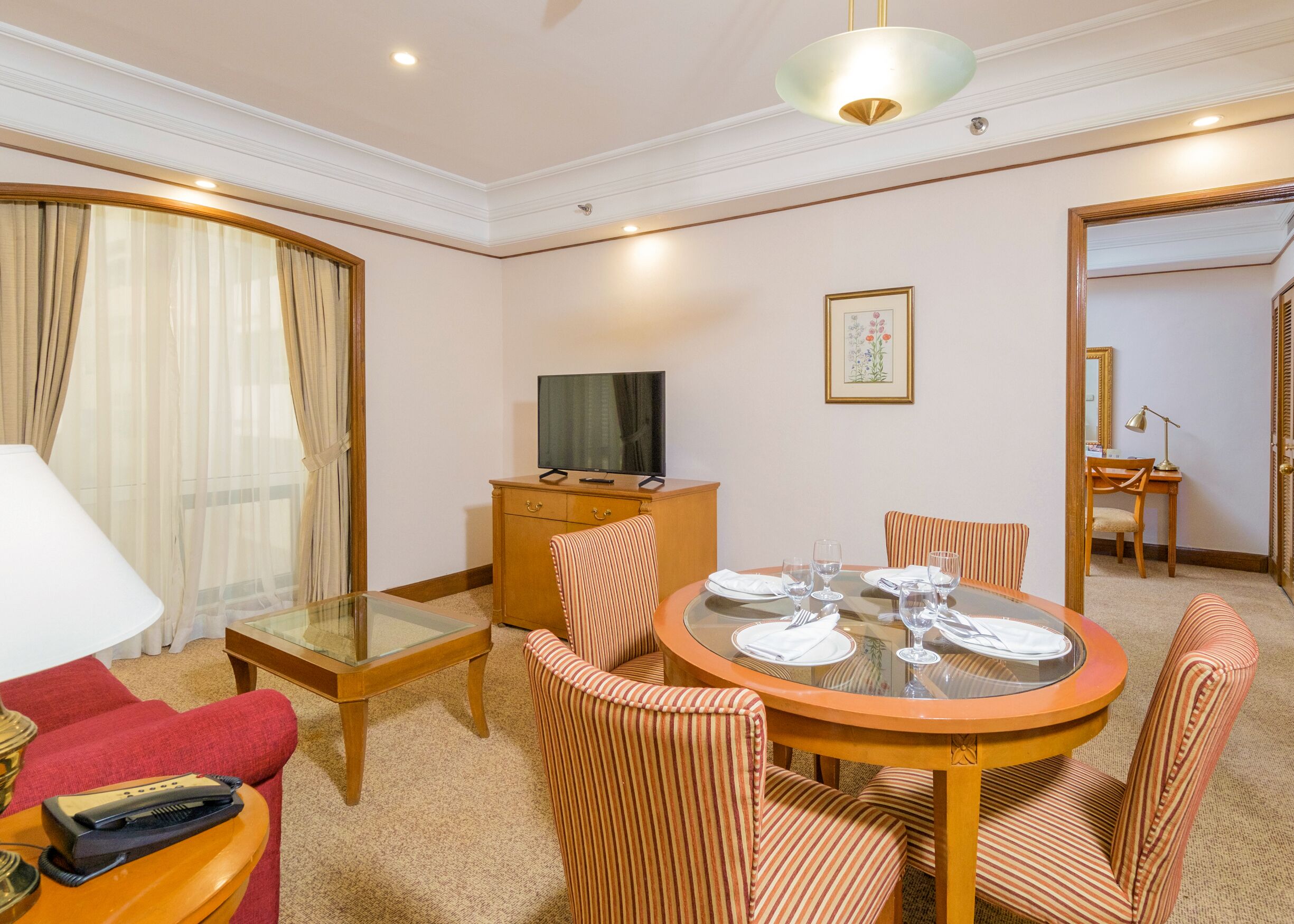 Une chambre d’hôtel avec une table à manger, des chaises à rayures, un téléviseur posé sur un meuble en bois, un canapé rouge et un grand miroir.