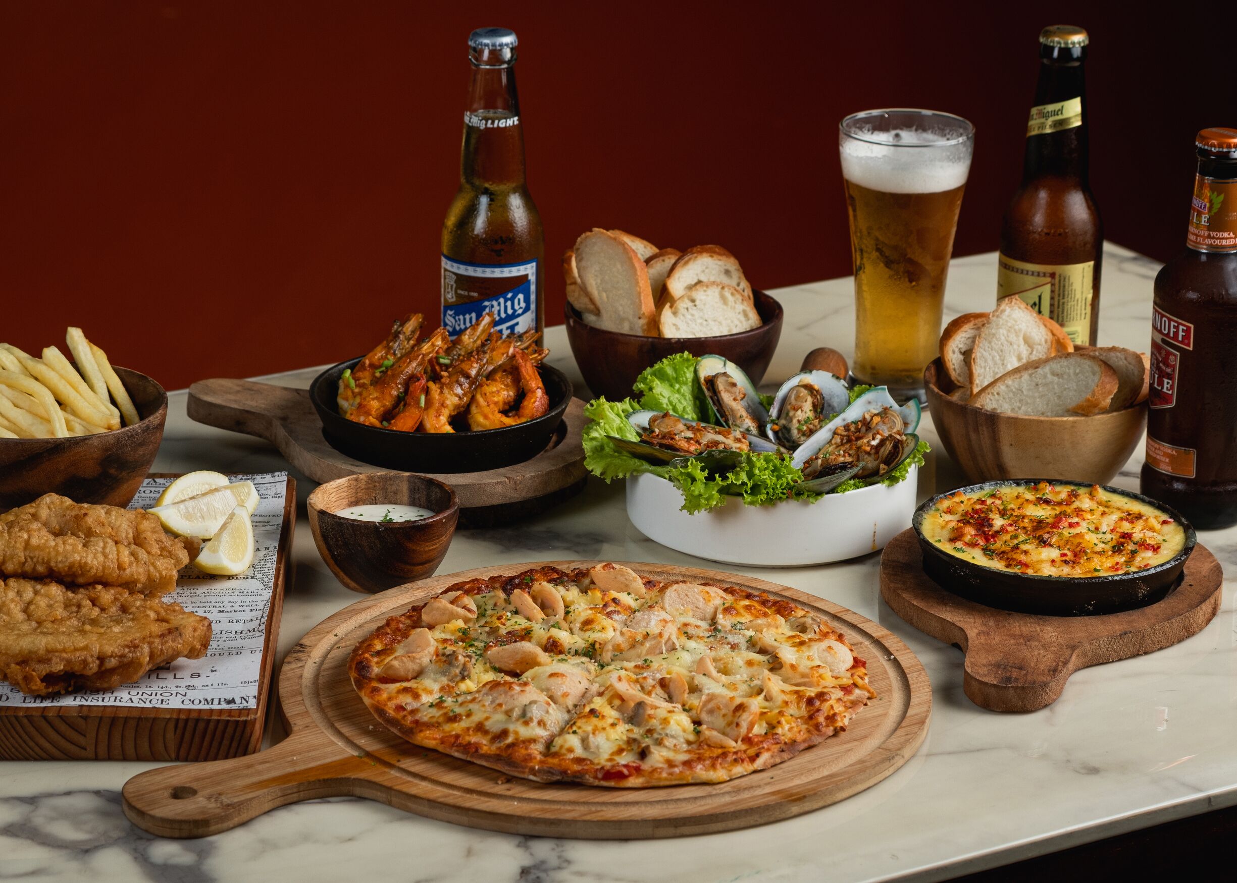 A variety of seafood dishes, including shrimp, mussels, and a seafood pizza, along with several bottles of beer and bread.