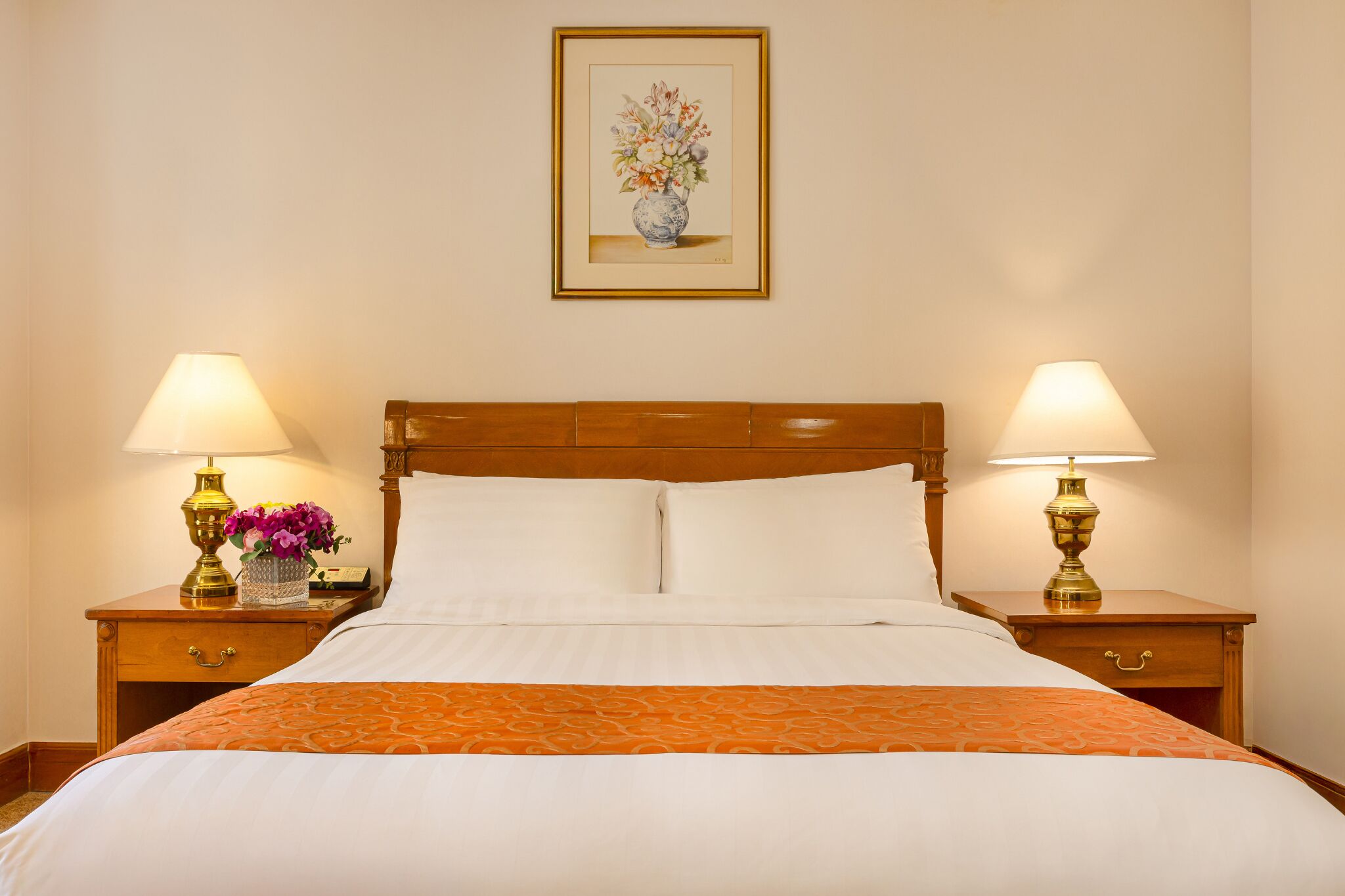 Una habitación de hotel con una cama de madera, dos mesitas de noche con lámparas, una obra de arte floral enmarcada en la pared y un jarrón con flores sobre una de las mesitas.