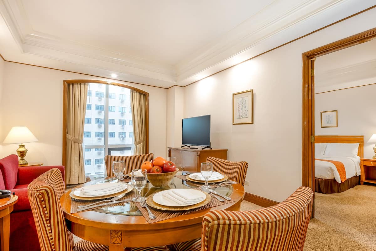 A hotel room with a dining table set for four, a television, a bed with a white comforter, and a window with curtains.
