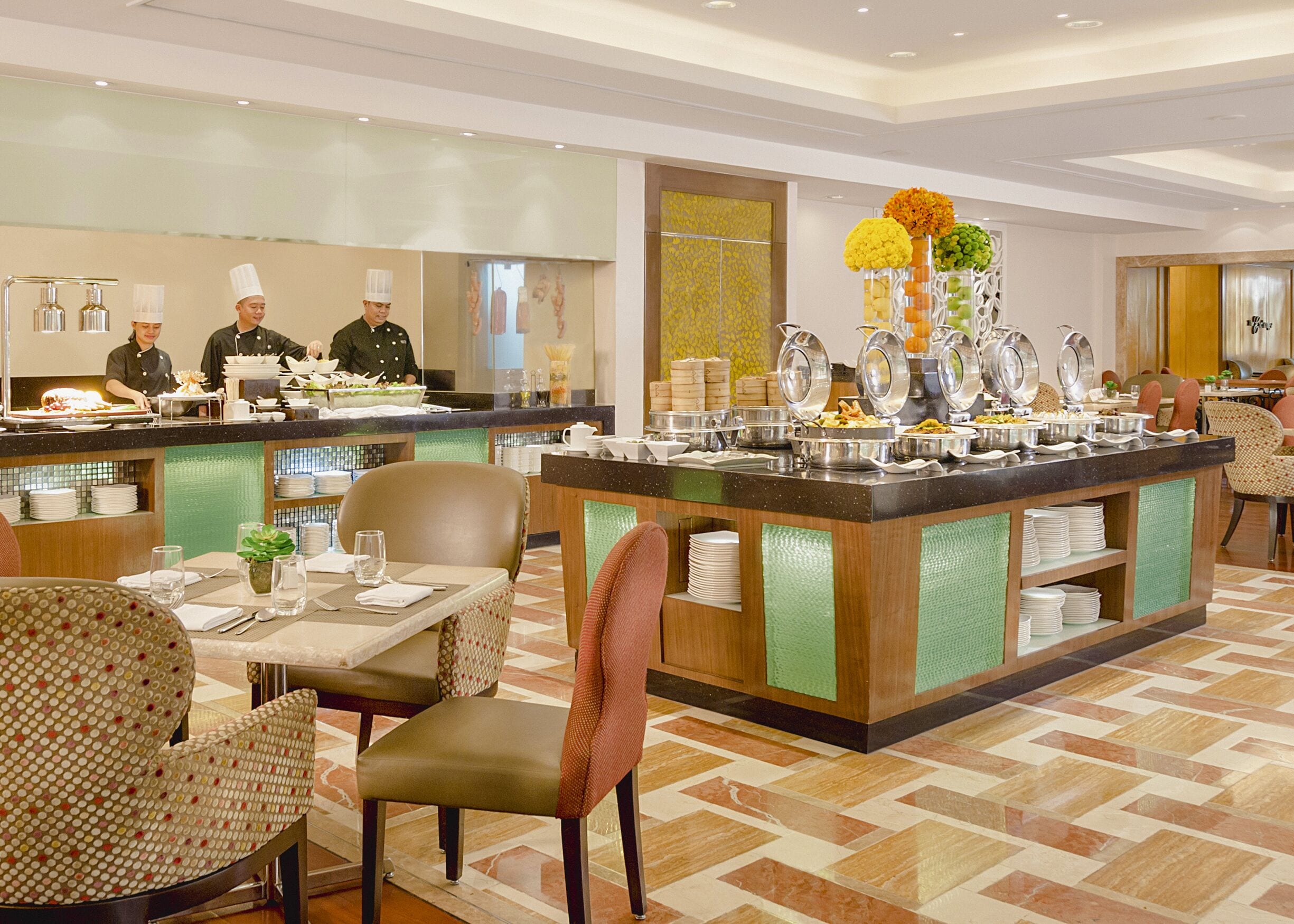 A hotel restaurant with a buffet setup, multiple dining tables, and a kitchen area with chefs.