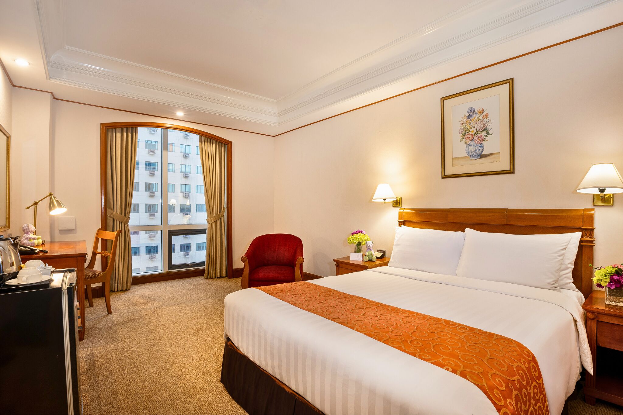 A hotel room with a large bed, a desk with a lamp, a chair, a window with curtains, and a painting on the wall.