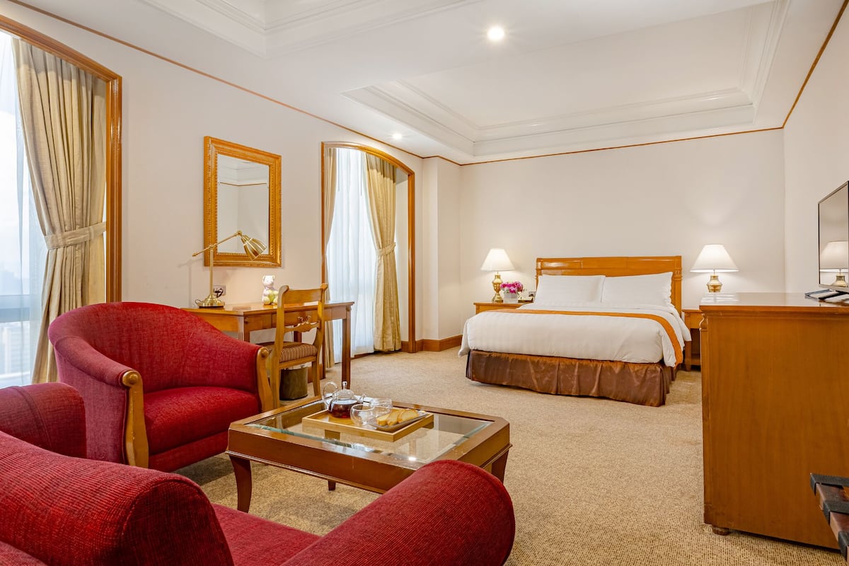 A hotel room with a bed, two red armchairs, a wooden desk, a mirror, and a view of the city.