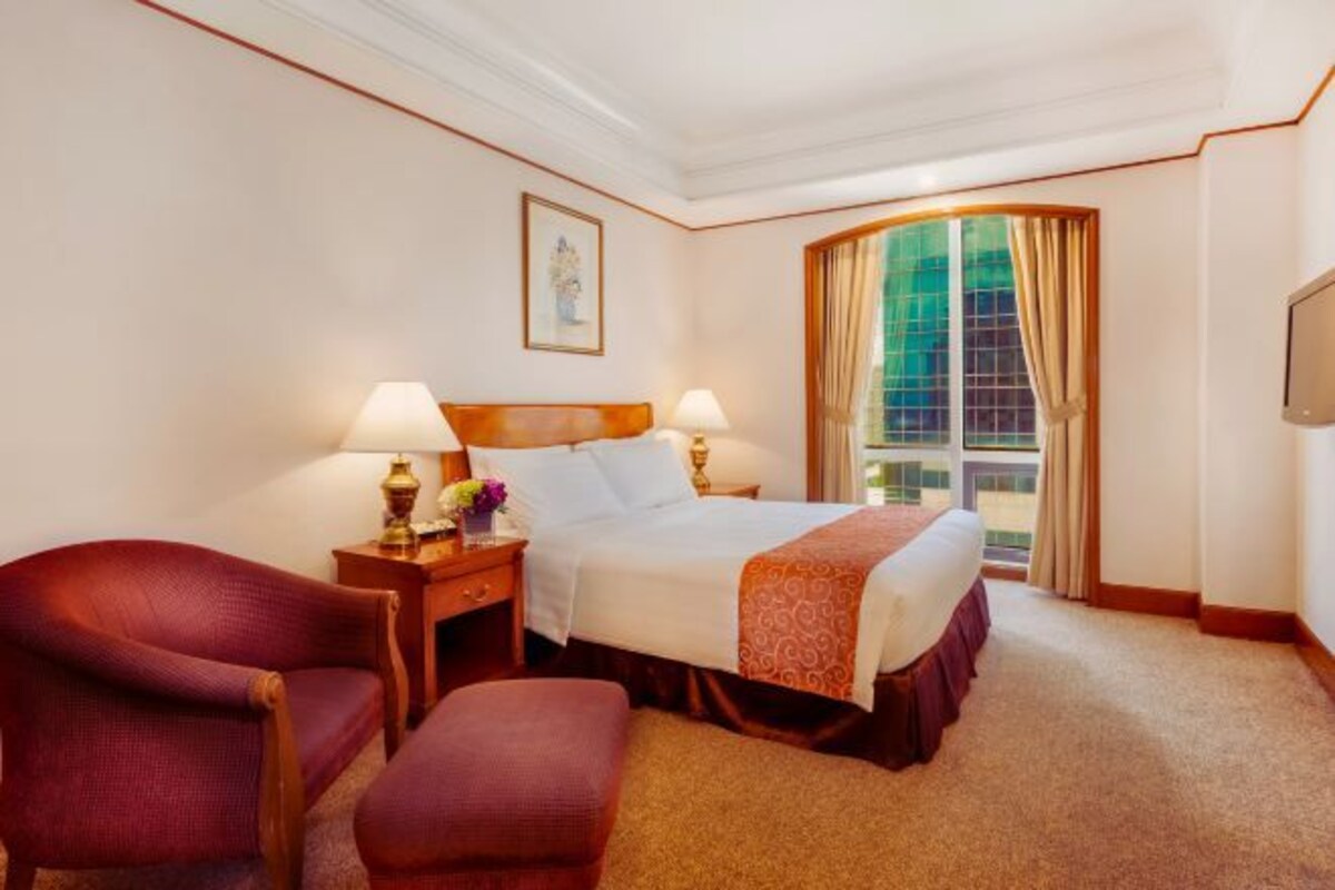 A hotel room with a large bed, two bedside tables with lamps, a red armchair, a footstool, and a large window with curtains.