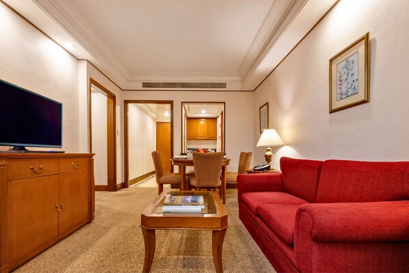 A hotel room with a red sofa, wooden cabinet, flat-screen TV, dining table with chairs, and a framed picture on the wall.