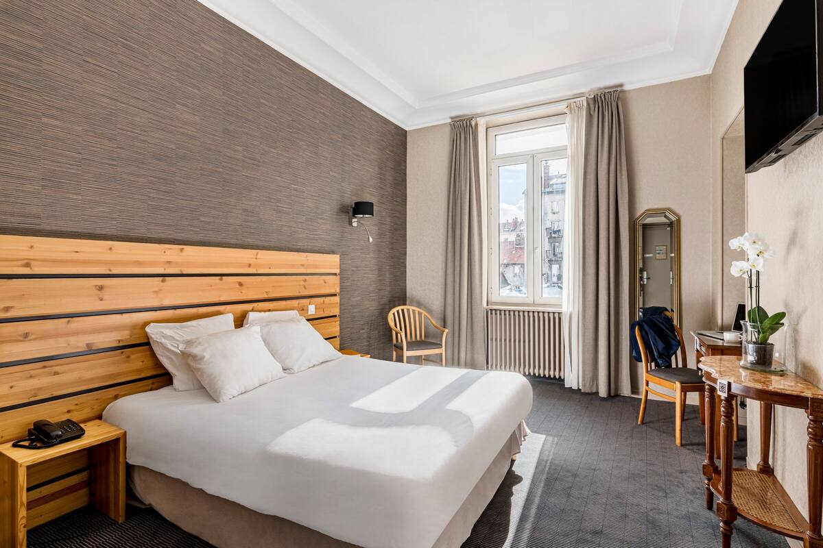 Una habitación de hotel con una cama grande, cabecero de madera, una mesita de noche con un teléfono, una silla, una pequeña mesa con una planta y un espejo en la pared.