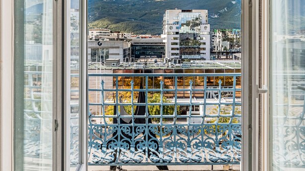 Un balcón con barandilla de metal, vista a edificios y montañas al fondo.