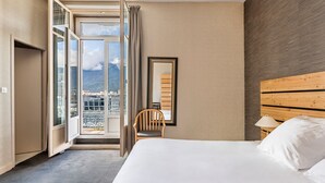 Habitación de hotel con una cama grande, cabecera de madera, una silla, mesita de noche con lámpara y vistas a un paisaje urbano a través de un ventanal amplio.