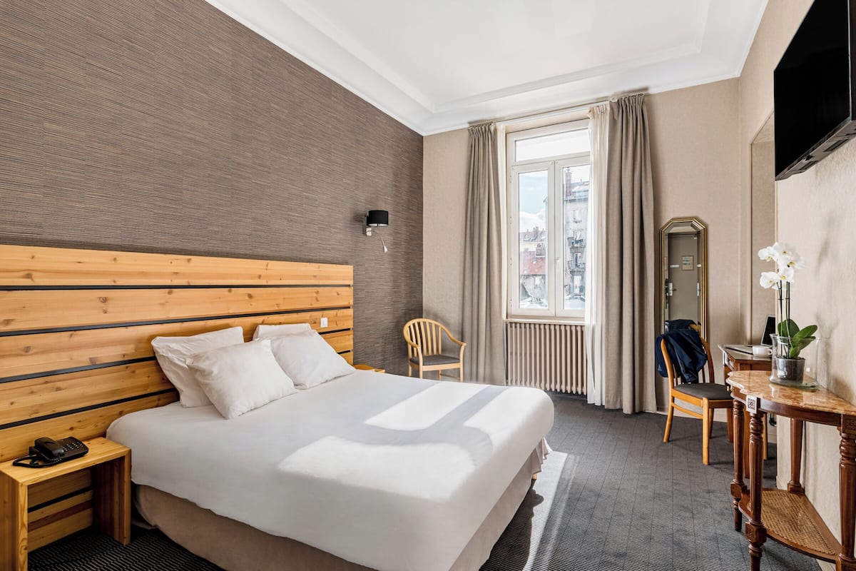 Camera d'albergo con un letto grande, una testiera in legno, un comodino con un telefono, una sedia, un tavolino con una pianta e uno specchio a parete.