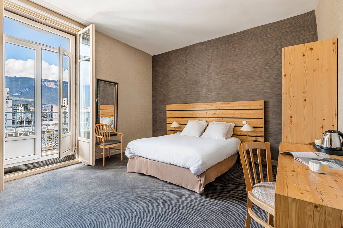 Habitación de hotel con una cama grande, cabecera de madera, un escritorio con silla, ventana con vista a las montañas y un balcón.