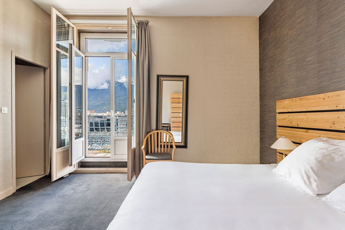 Camera d'albergo con un letto grande, una testiera in legno, una sedia, un comodino con una lampada e vista sulla città attraverso un'ampia finestra.