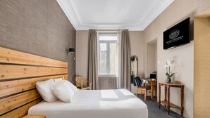 Habitación de hotel con una cama grande, un cabecero de madera, un escritorio con una silla, un televisor y una ventana con cortinas.