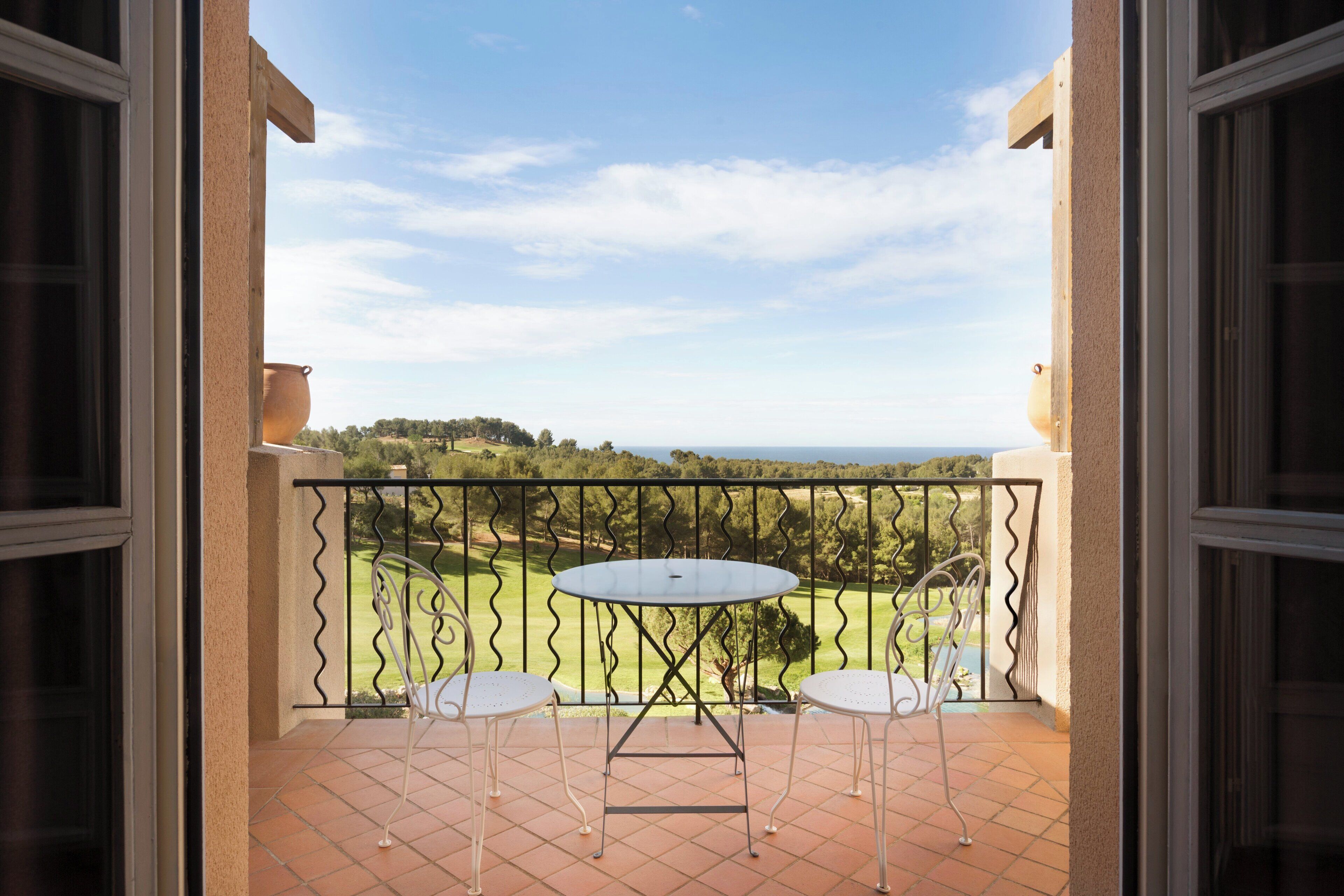 A balcony with a table and two chairs, overlooking a green landscape and a body of water.