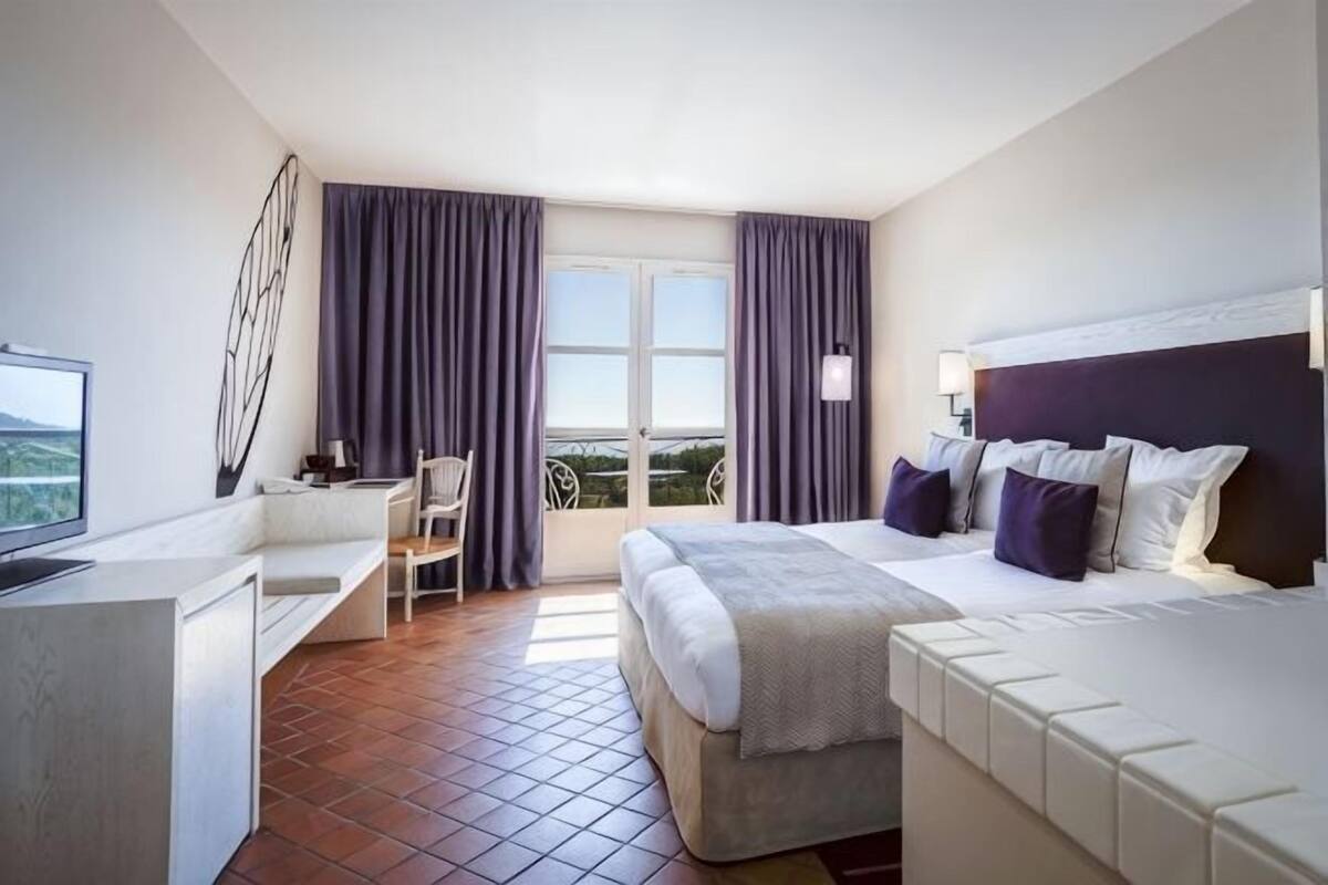 A hotel room with a bed, a desk, a chair, and a balcony with curtains.
