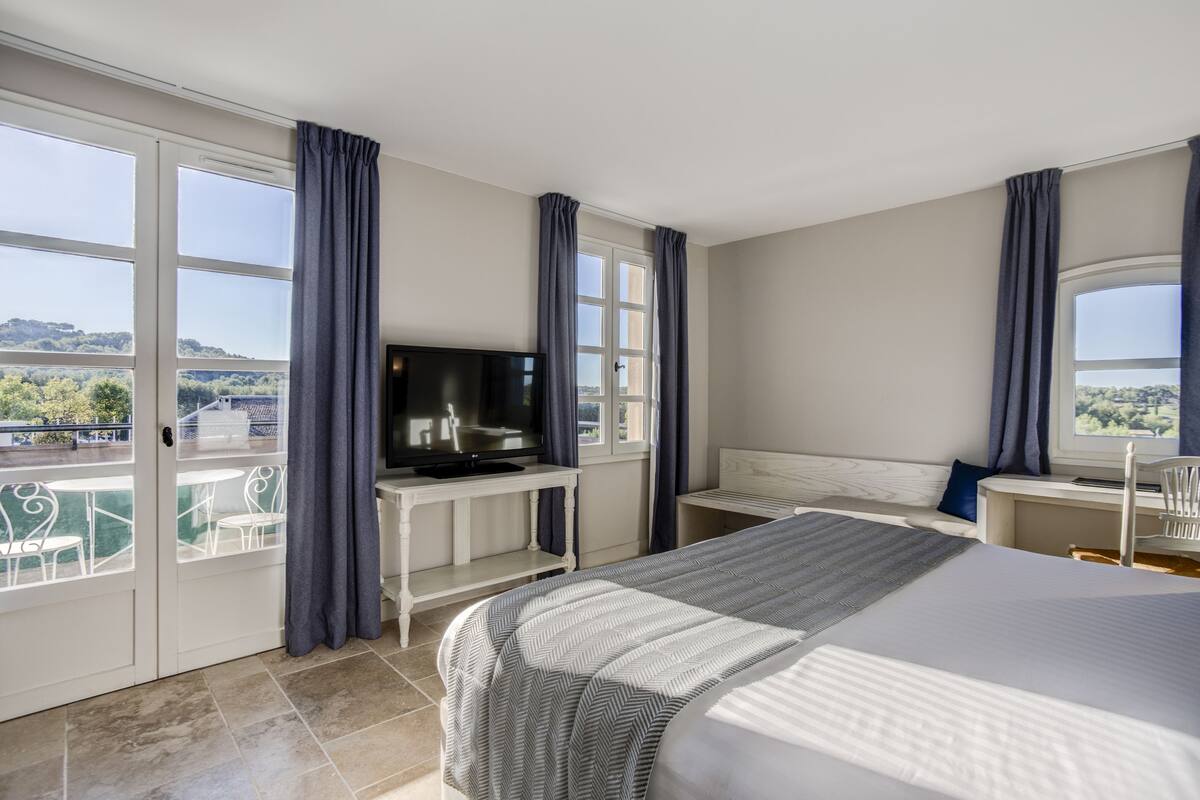 A hotel room with a bed, a desk with a TV, a balcony with chairs, and a view of the countryside.