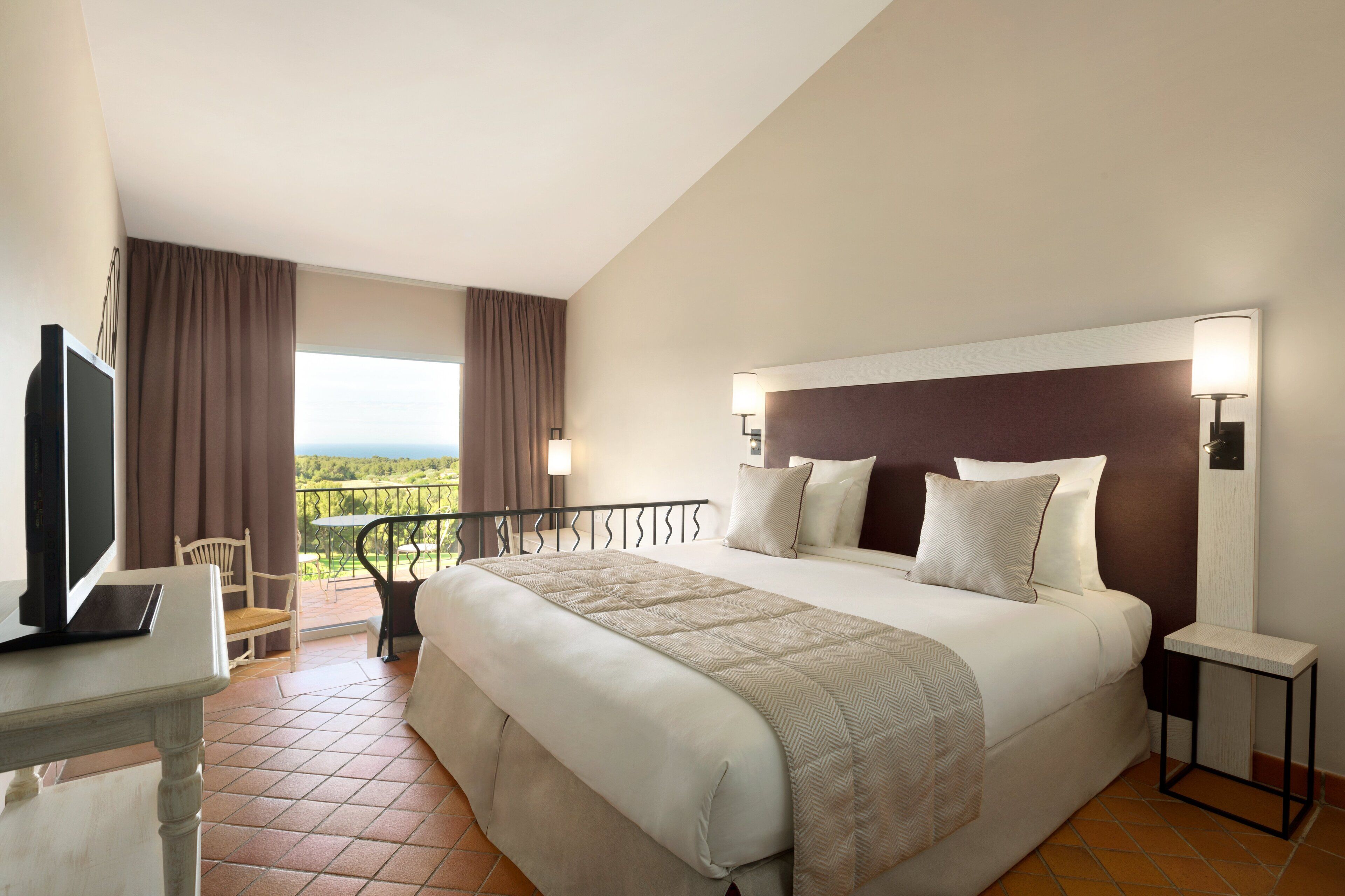 A hotel room with a large bed, a desk with a laptop, a TV, and a balcony with a view.