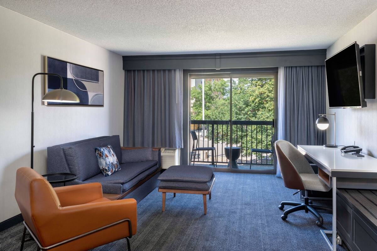 Ein Hotelzimmer mit einer Couch, Sesseln, Hocker, Schreibtisch und einem Balkon mit Blick auf Bäume.