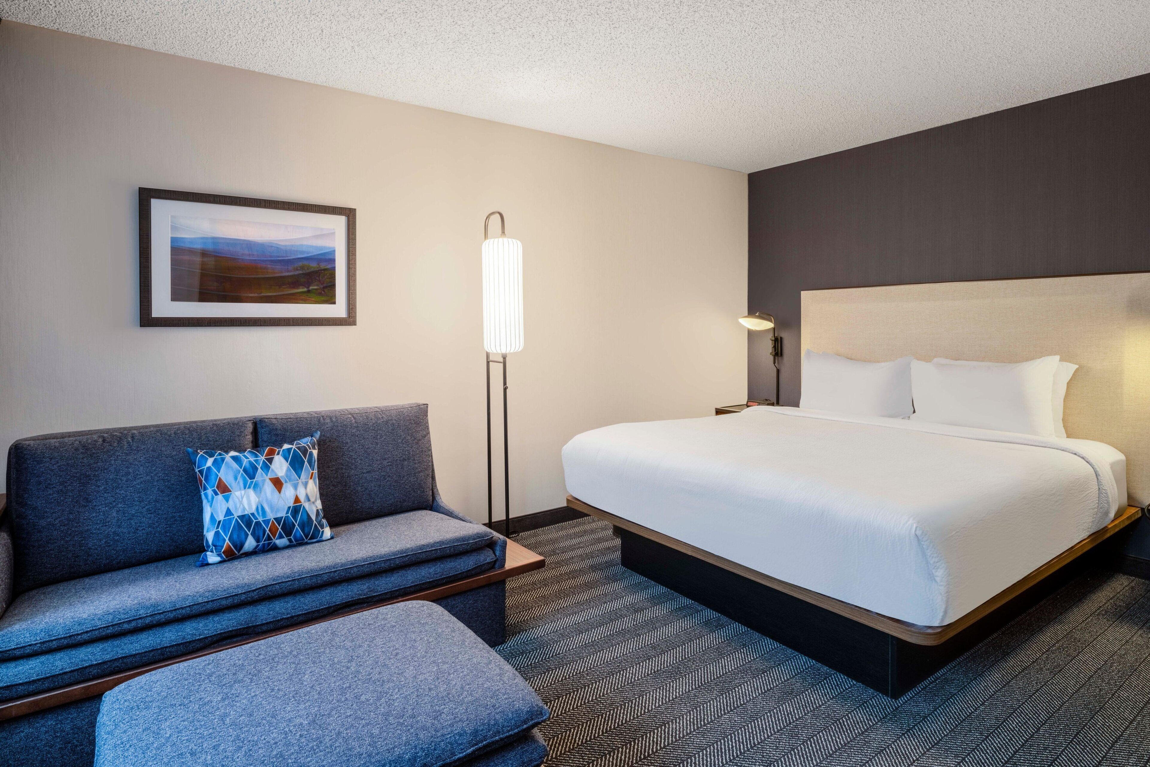 A hotel room with a bed, a sofa, a floor lamp, and a framed picture on the wall.