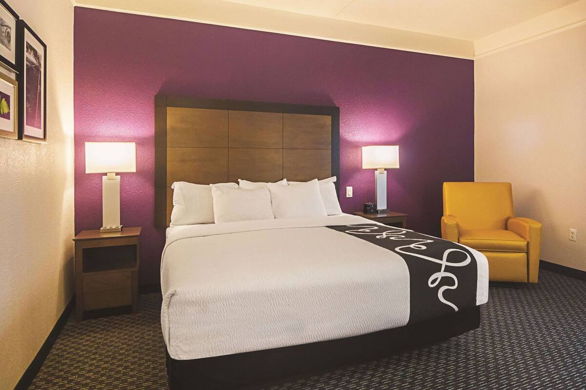 A hotel room with a bed, two bedside lamps, a chair, and a headboard.