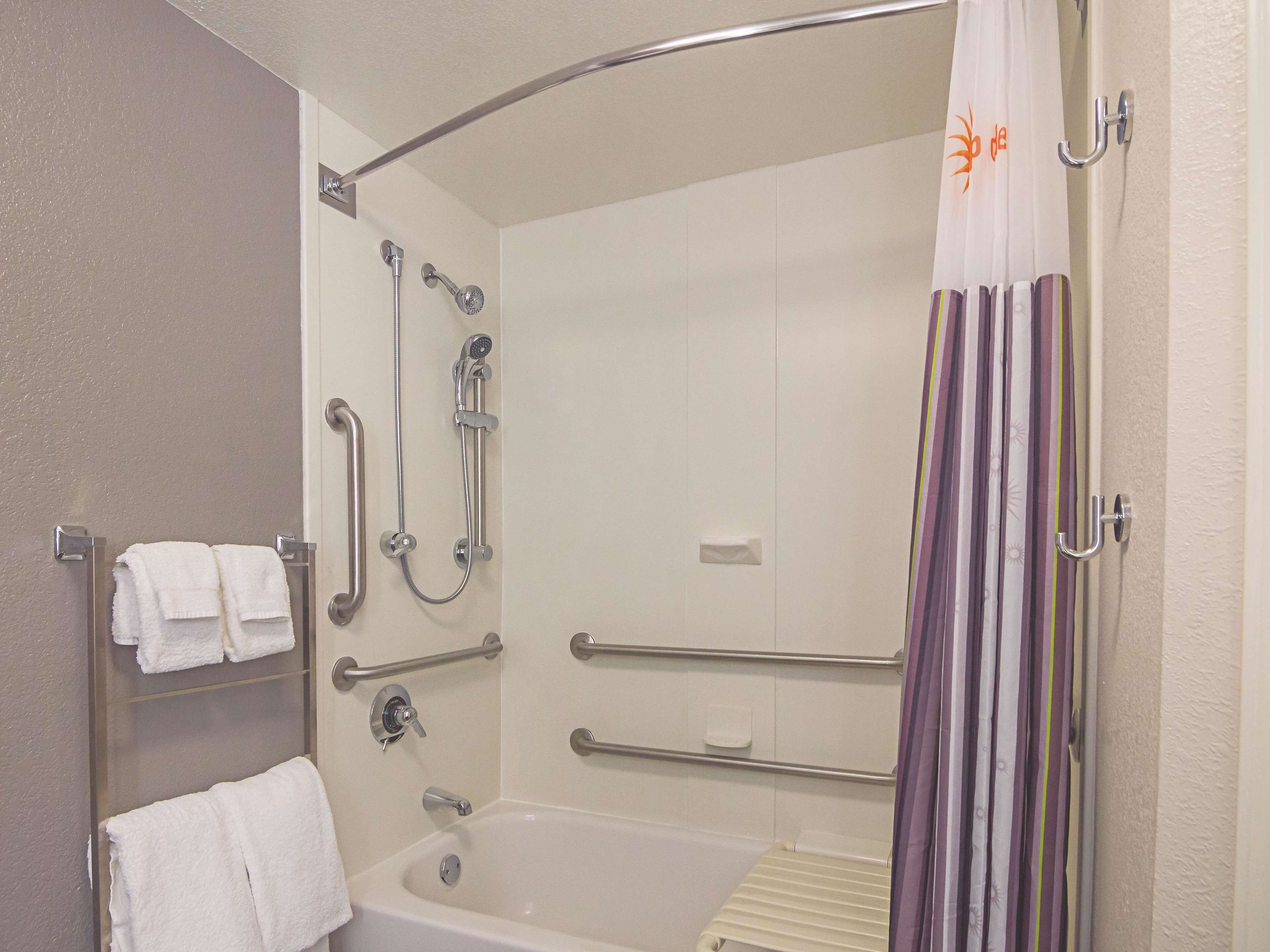 Baño con bañera, ducha con cabezal de ducha de mano, barras de apoyo y un perchero con toallas.
