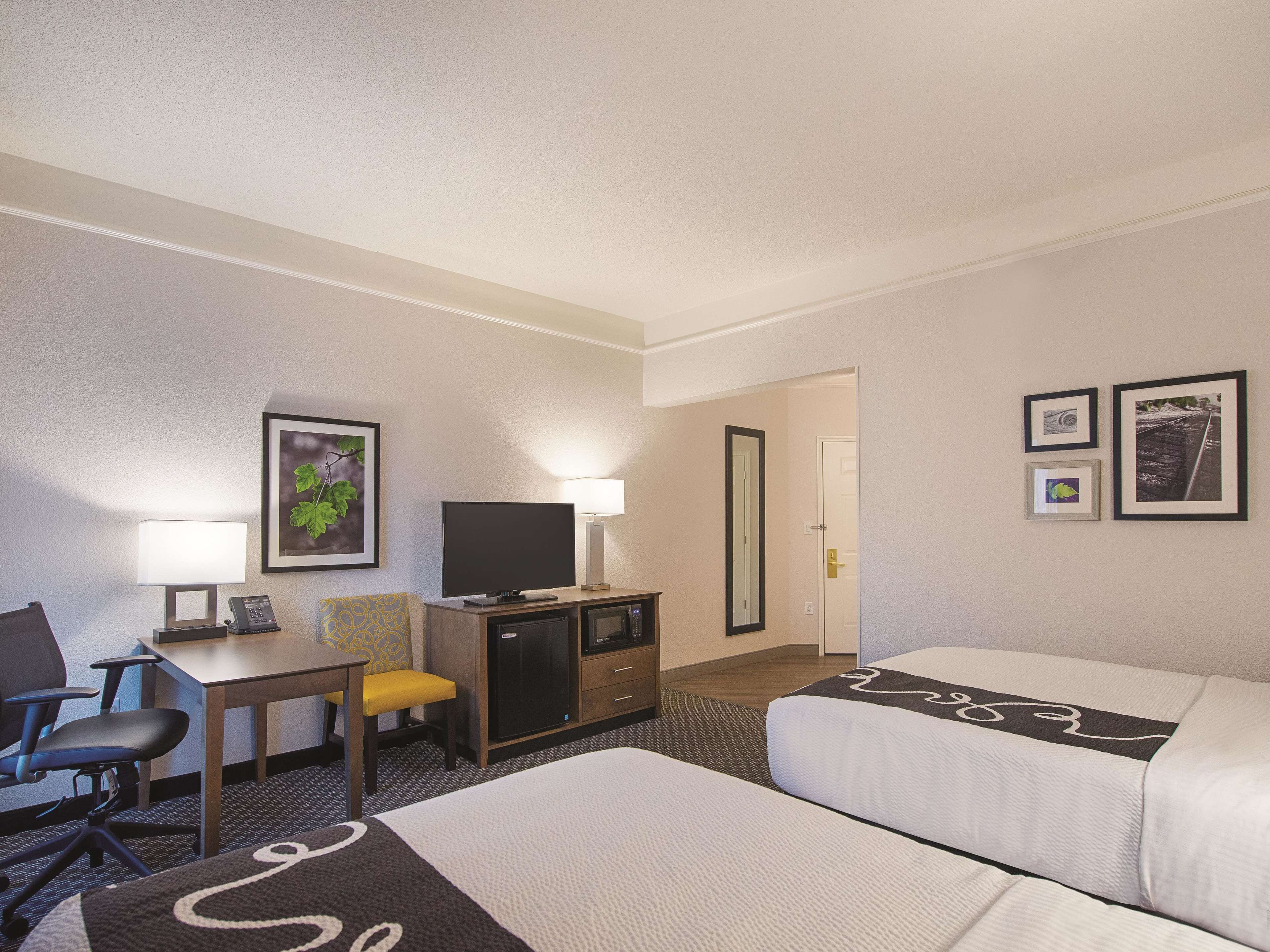 A hotel room with a bed, a desk with a chair, a television, a lamp, and framed pictures on the wall.