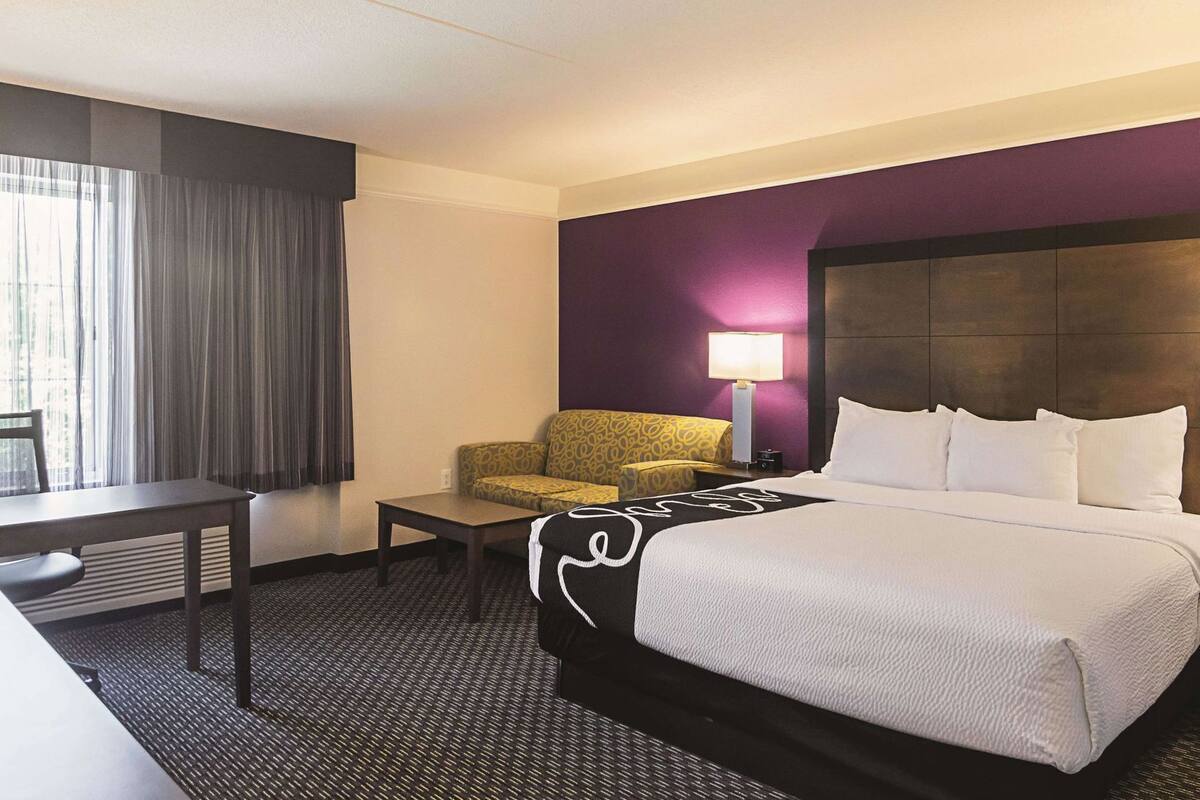 A hotel room with a bed, a desk, a chair, a lamp, and a window with curtains.