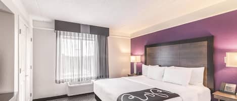 A hotel room with a large bed, a bedside table with a lamp, a window with sheer curtains, and a wall-mounted air conditioner.