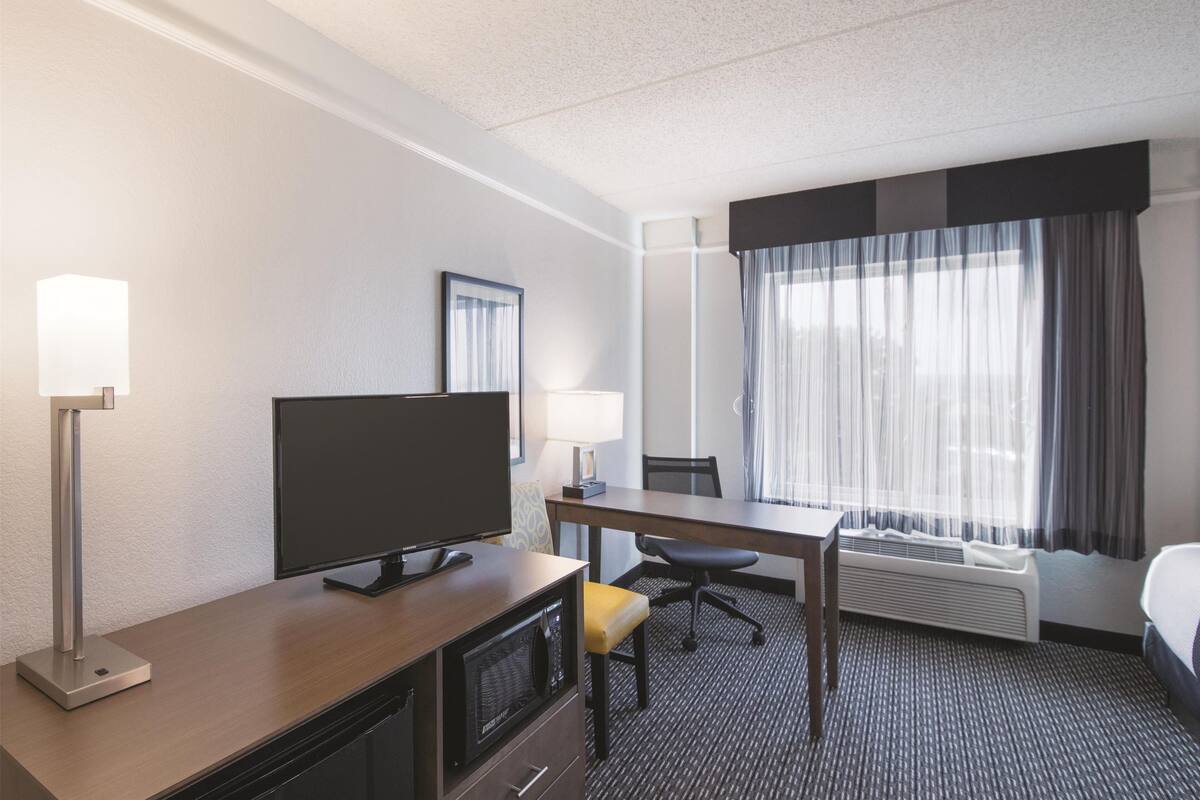 Une chambre d’hôtel équipée d’un téléviseur à écran plat, d’un bureau, d’une chaise, d’une lampe, d’un lit et d’une grande fenêtre avec des rideaux.