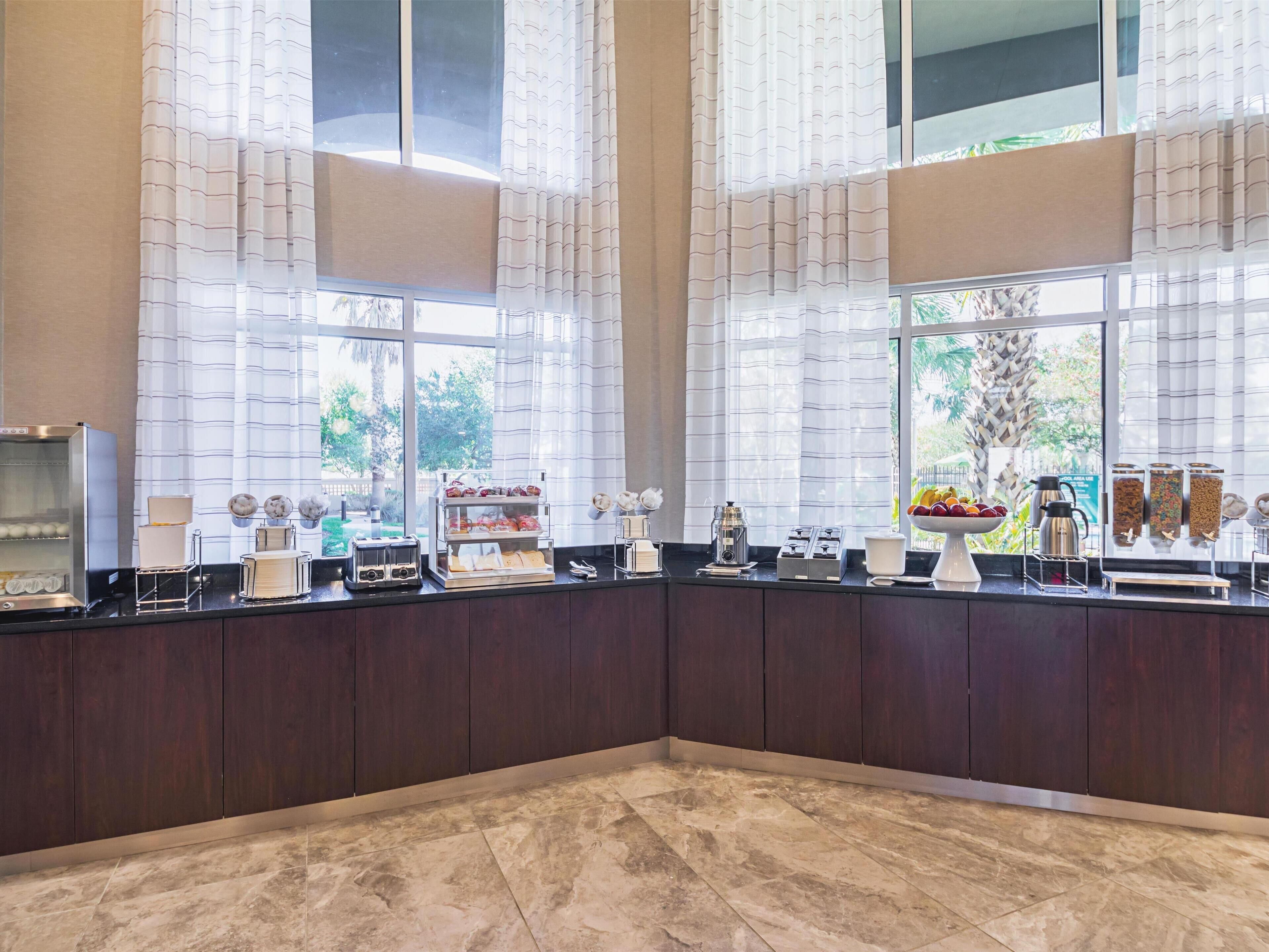 A modern kitchen with a marble floor, large windows with sheer curtains, and a variety of breakfast items displayed on the counter.